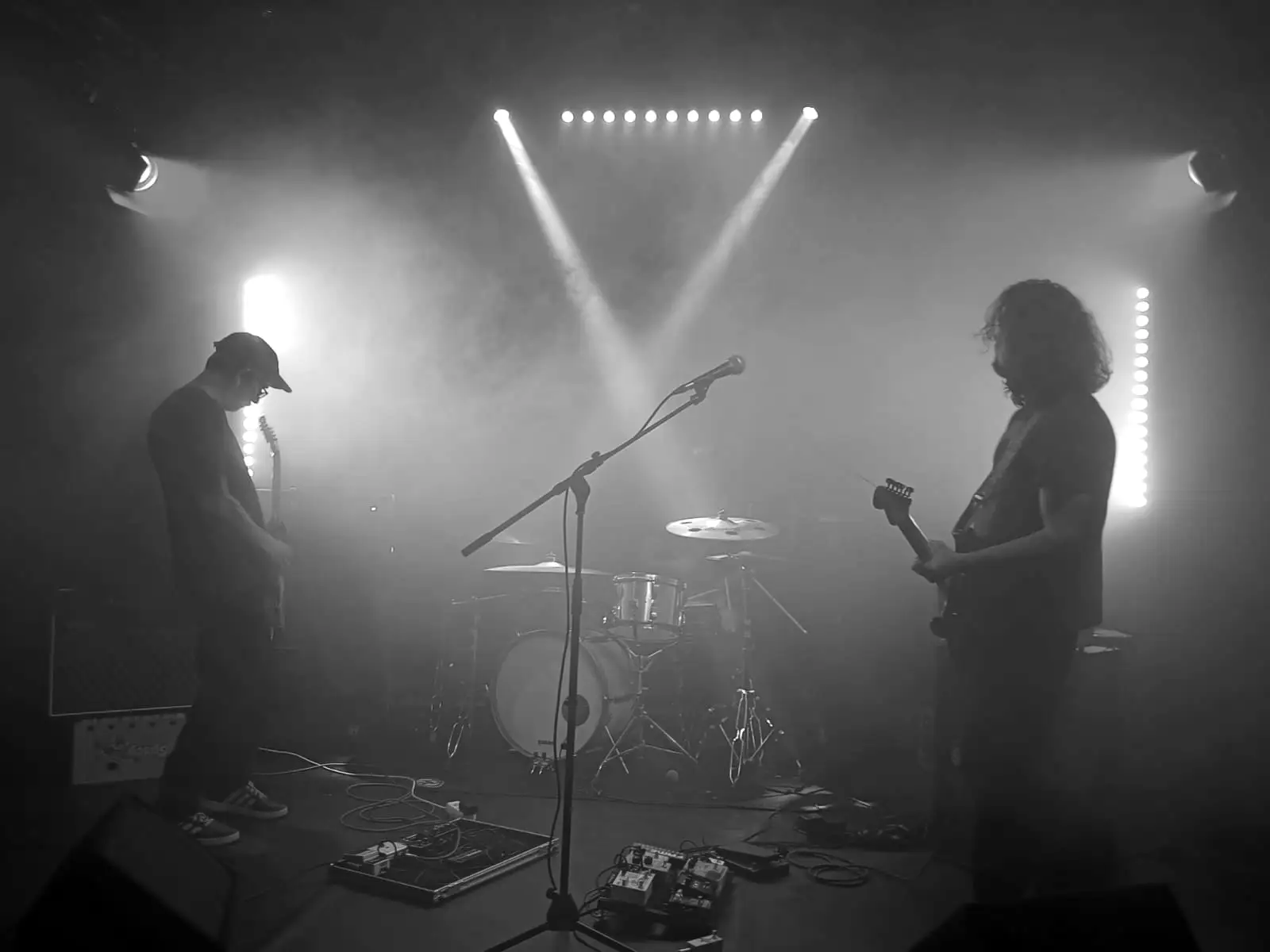 Guitarist and bassist facing each other, drum kit between them, light beams in the haze