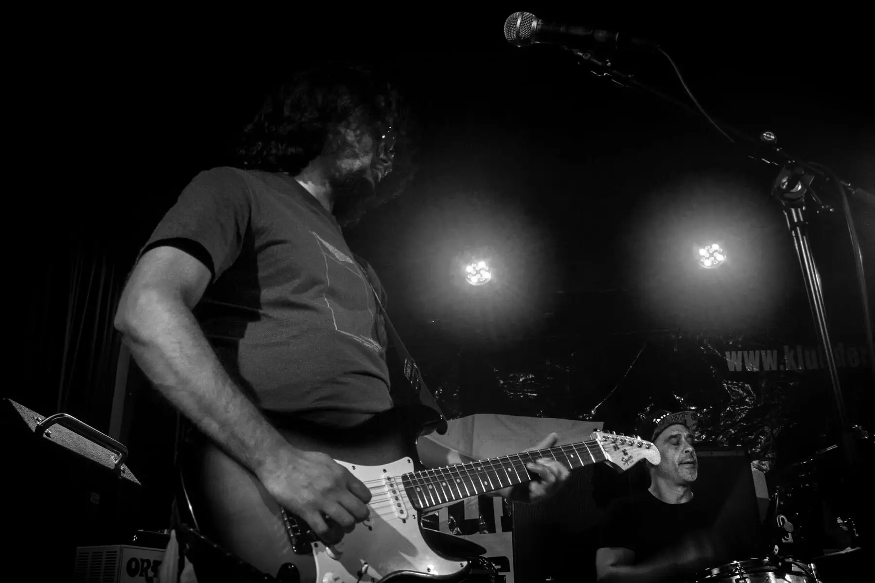 Guitarist and singing drummer between two glowing stage lights